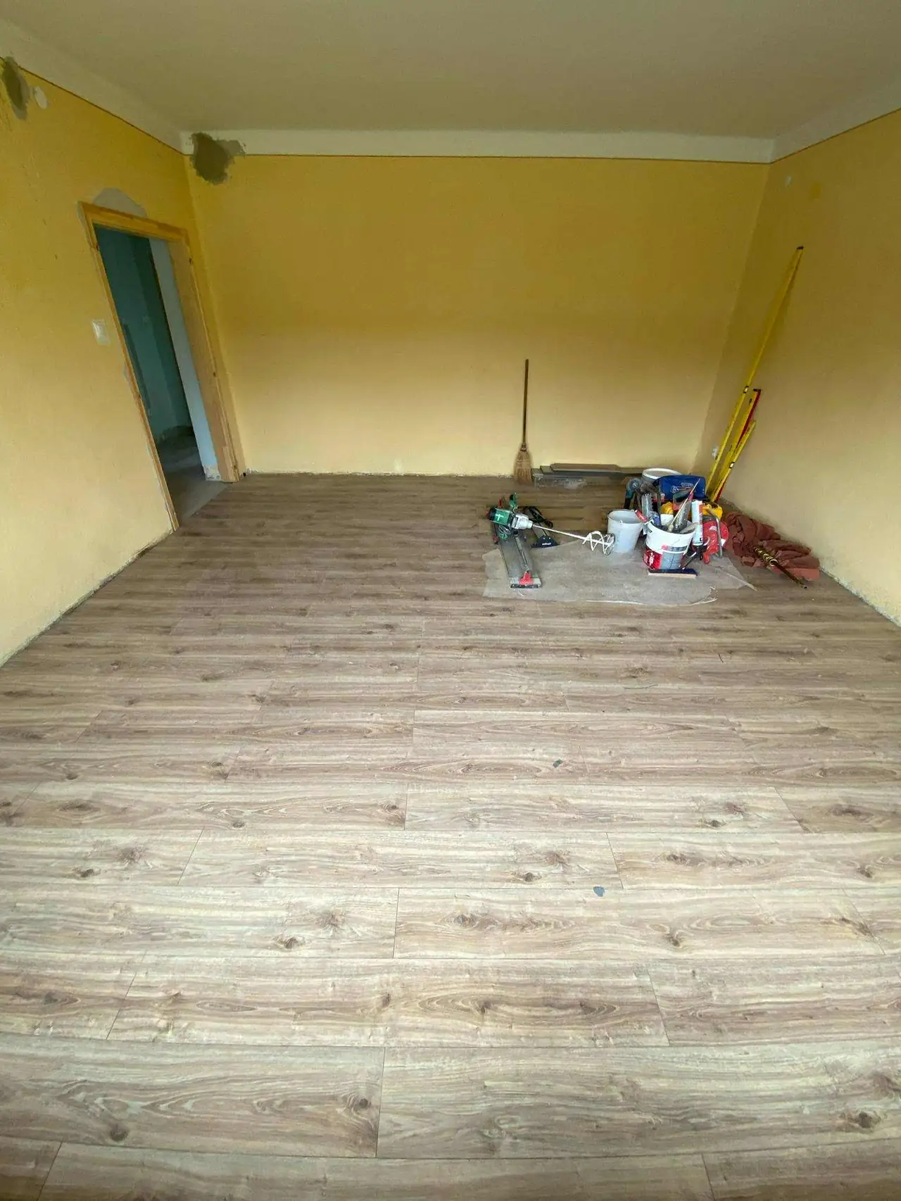 A képen egy felújítás alatt álló szoba látható, amelynek falai világos sárga színűek. A padló új, fa hatású burkolattal van ellátva. Az egyik sarokban takarítóeszközök és festési kellékek találhatók, míg a bejáratnál egy nyitott ajtó vezet egy másik helyiségbe. A Farkas-Konstrukt Kft. Generálkivitelezés szolgáltatásával készült felújítás Bük városában zajlik.