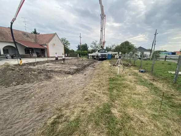 A képen a Farkas-Konstrukt Kft. földmunkát végez Győr városában. A háttérben egy épület látható, amely mellett munkások dolgoznak. Az építkezési területen nagy gépek, például betonpumpa és földmunkagépek találhatóak. A környező terület zöld fűvel borított, kerítés és fákkal van körülvéve. A generálkivitelezés szakszerű kivitelezése jól tükröződik a képen.