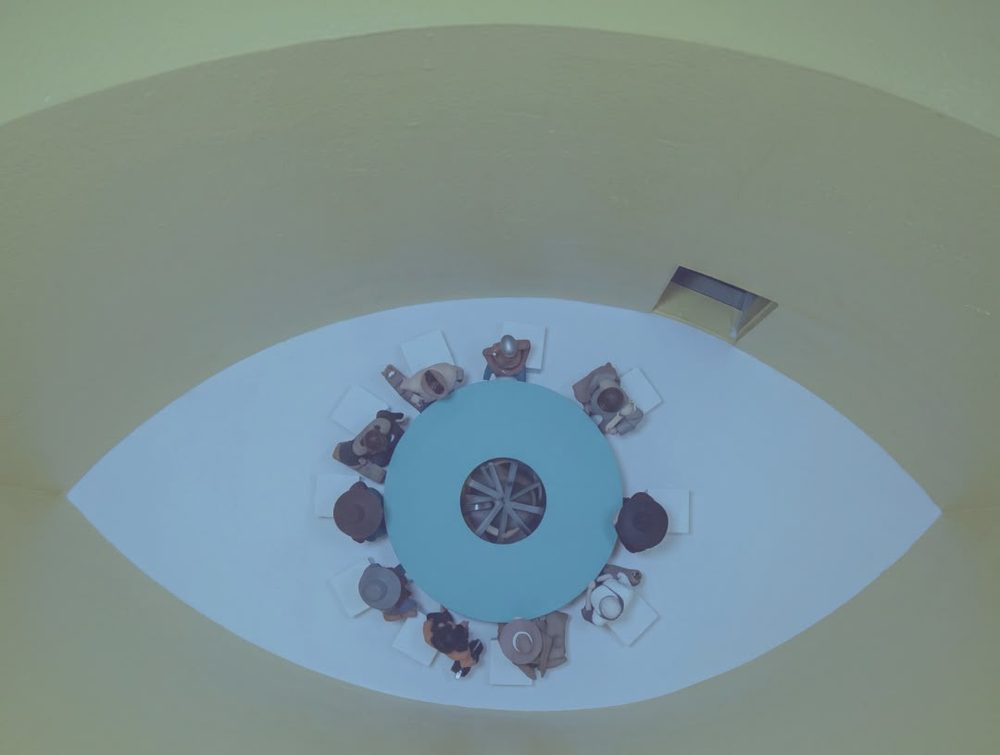 Mesa redonda con personas sentadas, vista desde arriba, dentro de la forma de un ojo