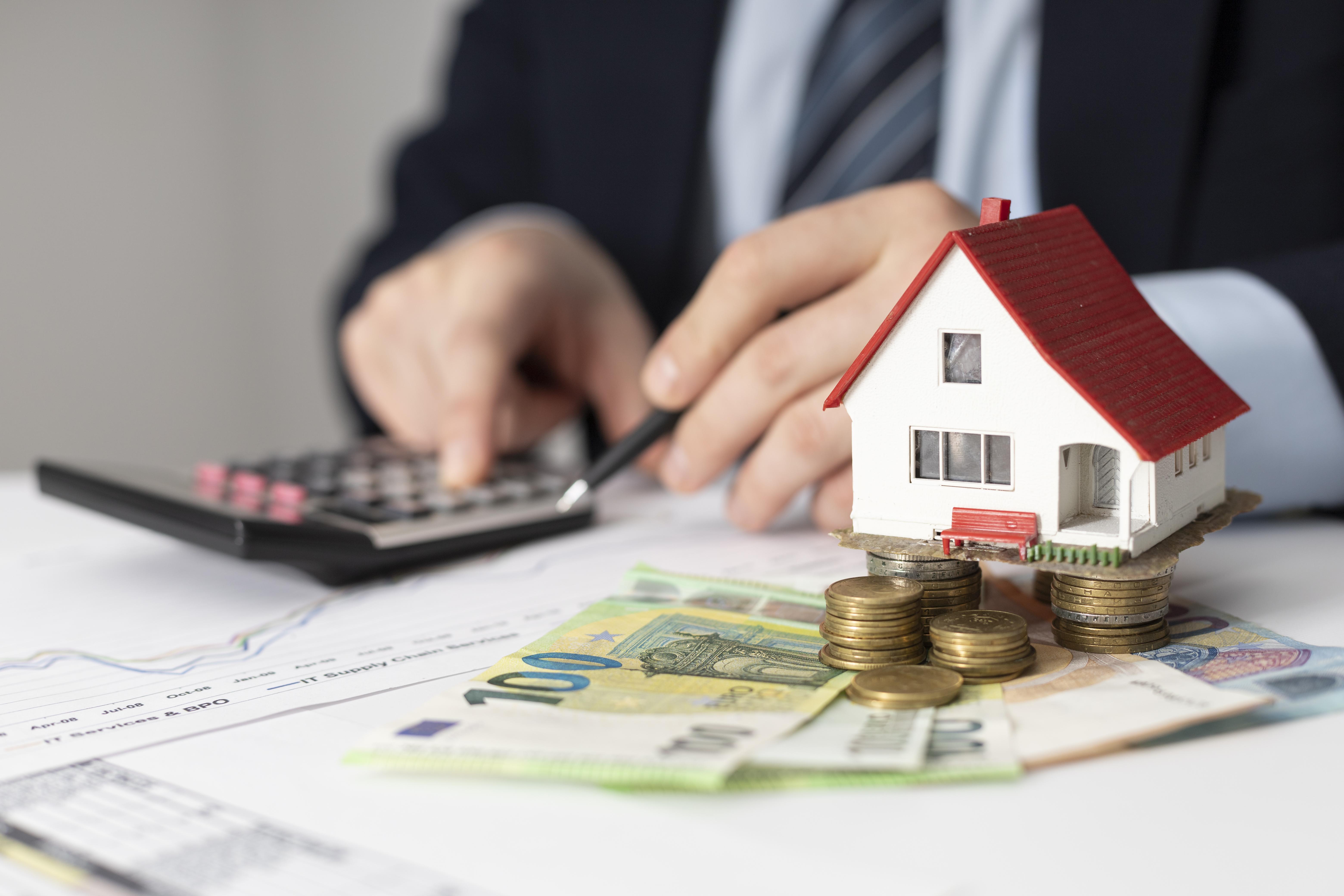 Real estate investment concept with a miniature house, coins, cash, calculator, and documents on a desk.