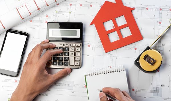  Hands using a calculator and writing notes on architectural blueprints with a red house cutout and measuring tape, representing home construction planning and budgeting.