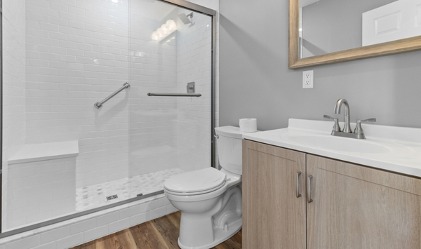 Compact bathroom with glass walk-in shower, built-in bench, grab bars, white toilet, wood-tone vanity, large mirror, and gray walls. Compact bathroom with glass walk-in shower, built-in bench, grab bars, white toilet, wood-tone vanity, large mirror, and gray walls.