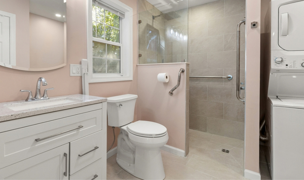 Small bathroom with white vanity, toilet, glass walk-in shower, grab bars, window, and stacked washer and dryer. Small bathroom with white vanity, toilet, glass walk-in shower, grab bars, window, and stacked washer and dryer.