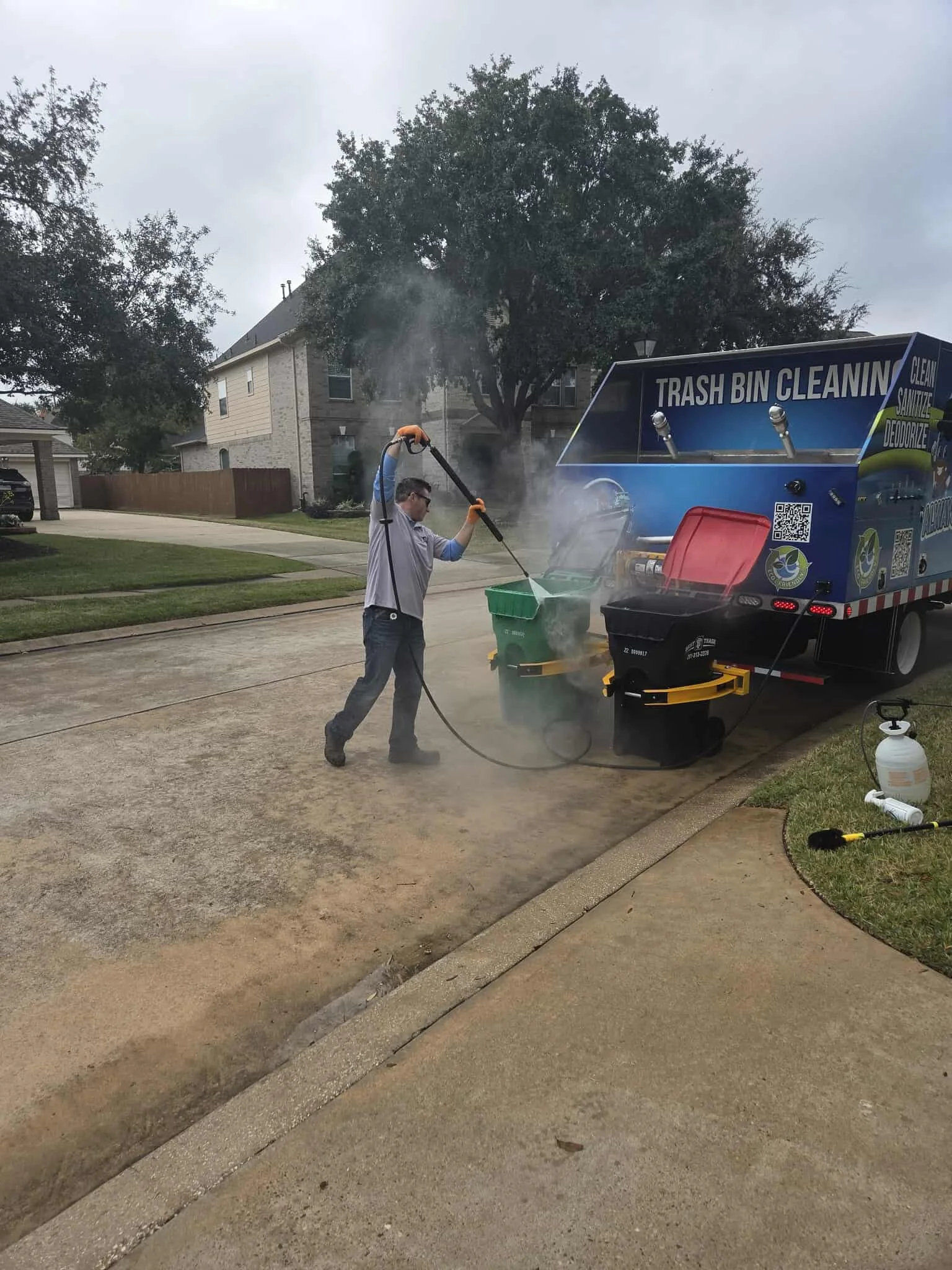 A man in safety glasses and gloves power washes two trash bins against the side of a sanitation truck.