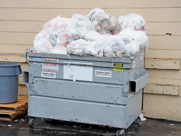 An overflowing gray dumpster with trash bags piled high, against a beige wall and wet ground.