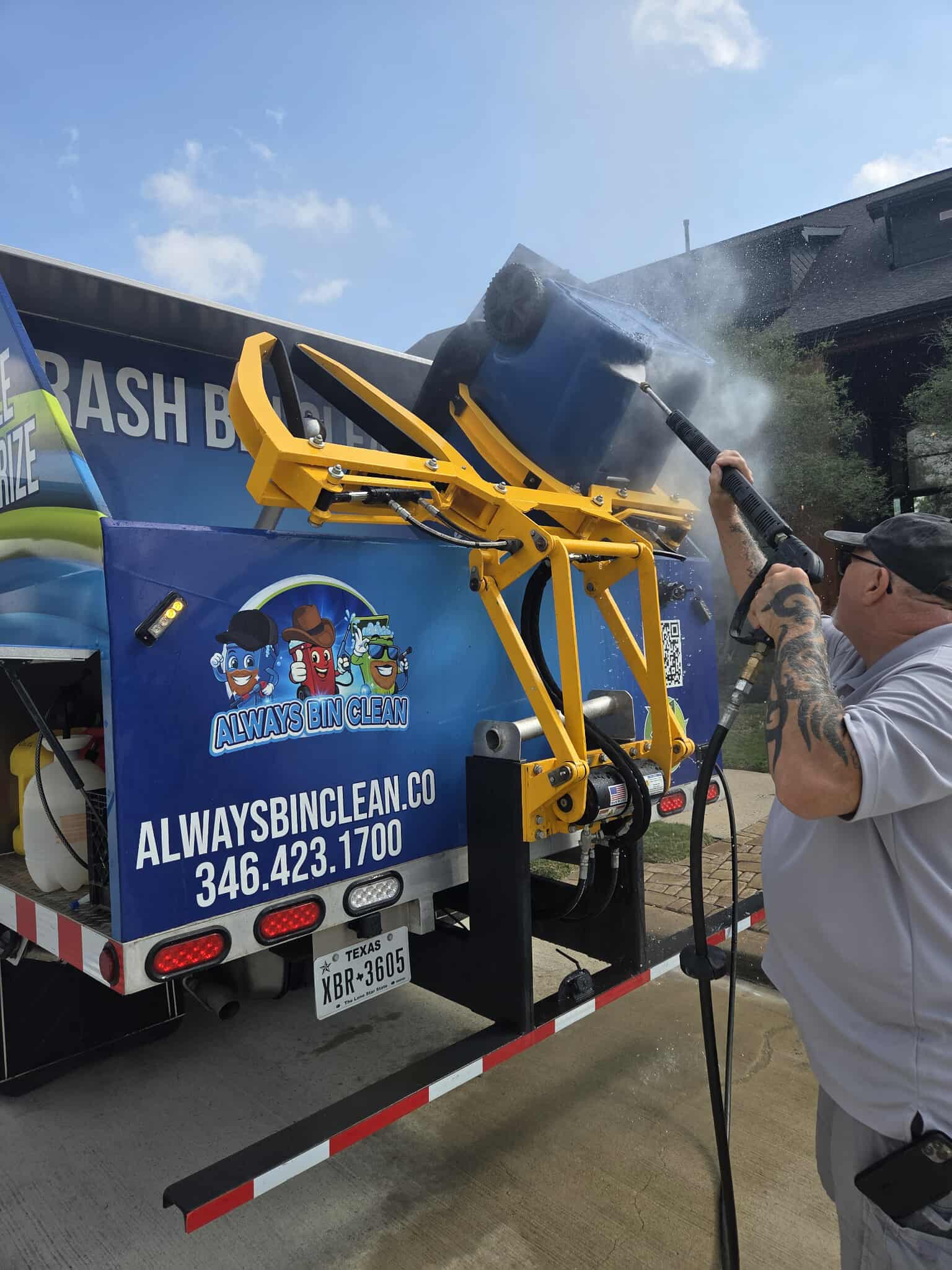 A man in orange gloves steam cleaning a green and black wheelie bin on a driveway next to a sanitation truck.