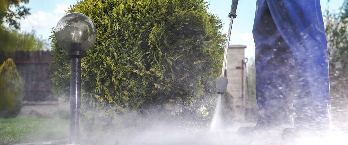 A person in work pants power washing a concrete patio next to green shrubs and rocks on a sunny day.