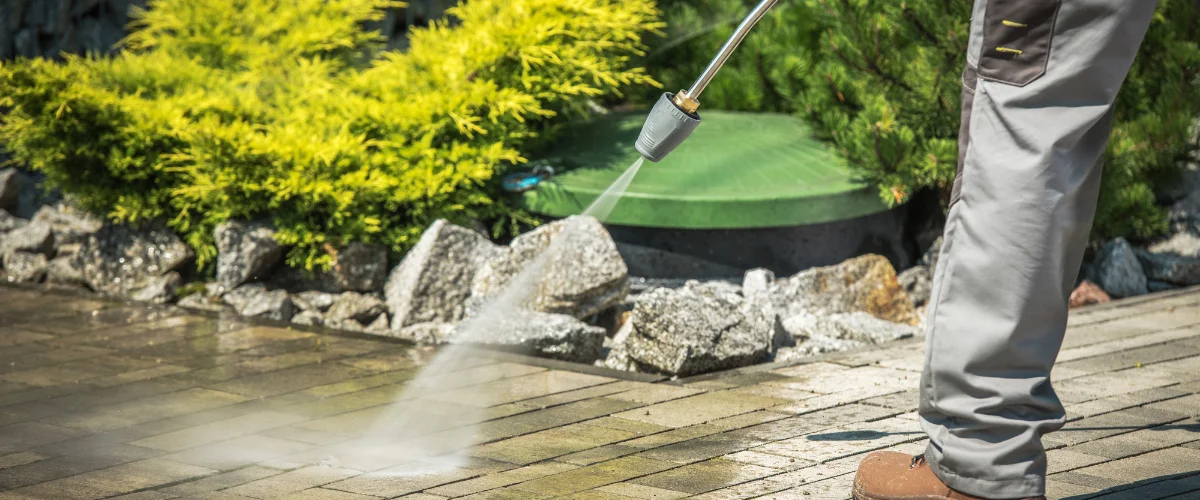 A person in blue overalls power washing a patio, with a green bush and garden lamp in the background.
