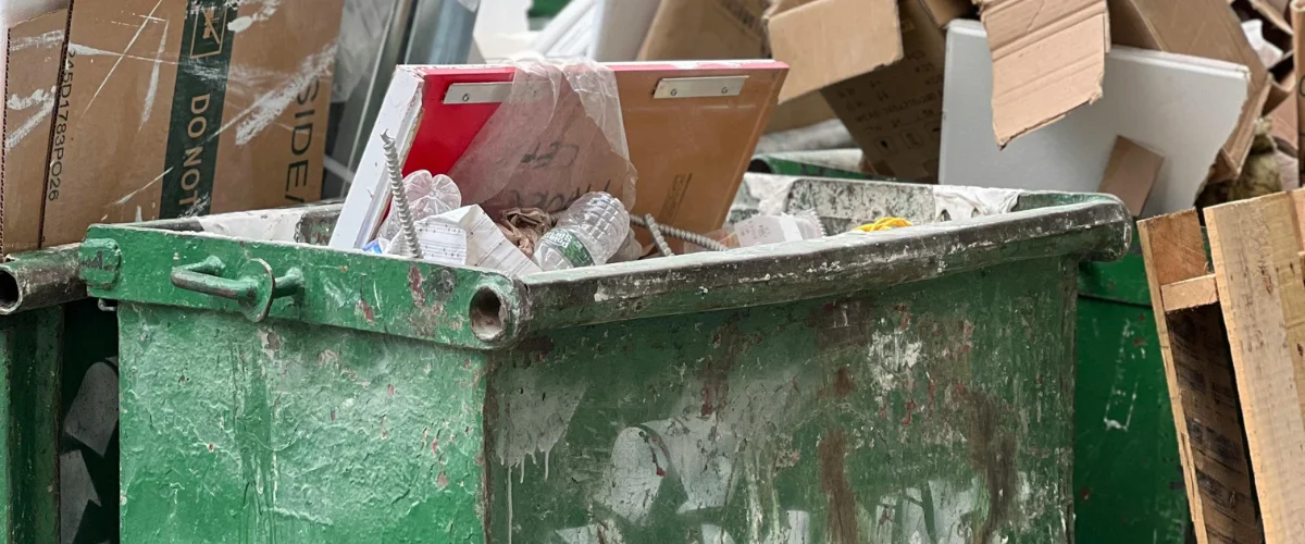 A close-up view of a green, paint-splattered dumpster overflowing with cardboard boxes and various waste items.