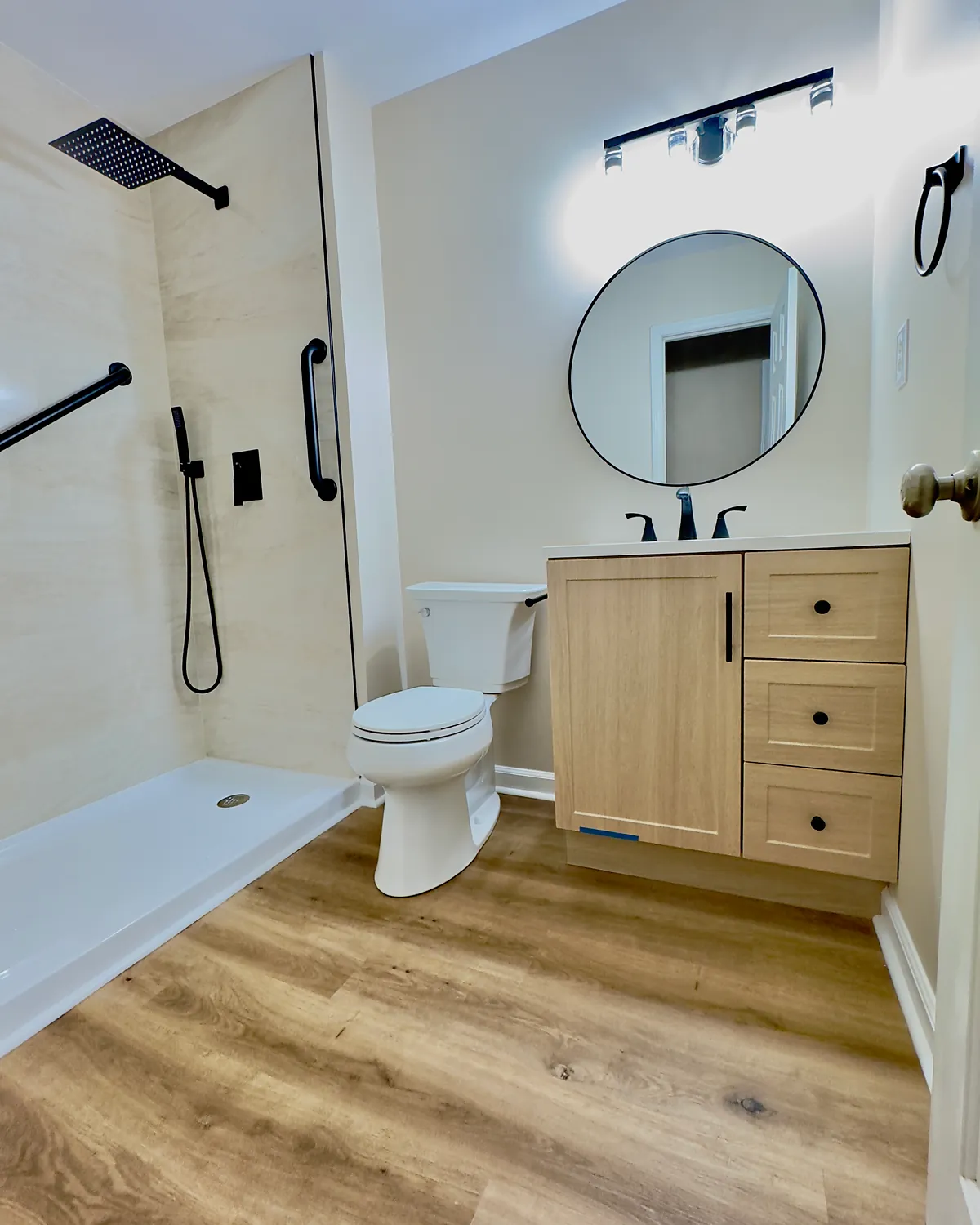 Walk-in shower with grab bars, rain shower head, and a light oak vanity with a round mirror