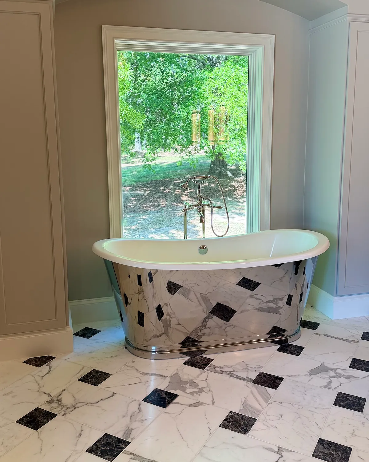 Polished metal freestanding tub under an arched window with a marble and black diamond tile floor