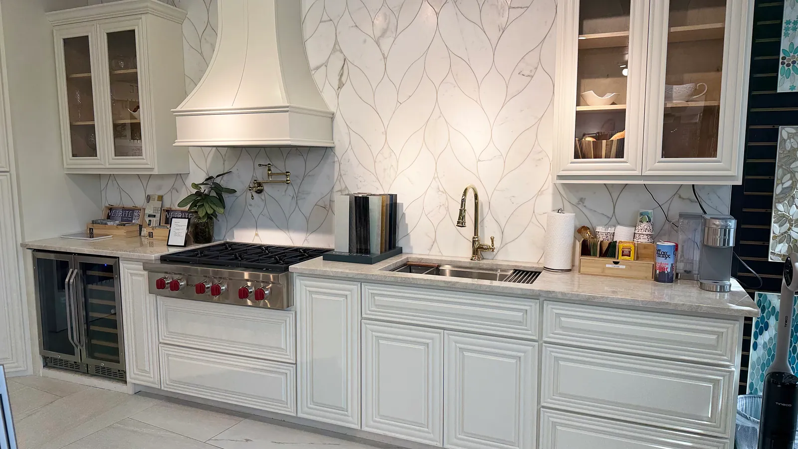 White kitchen with a custom range hood, leaf-pattern marble backsplash, and wine cooler