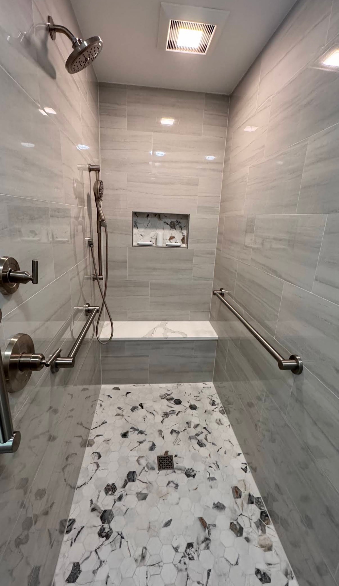 Gray-tiled shower with a built-in bench, grab bars, handheld shower and mosaic floor.