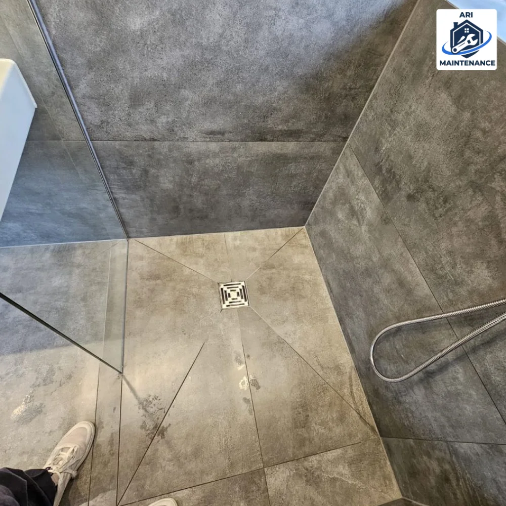 Walk-in shower close-up with linear drain — worker boot just visible at edge of frame.