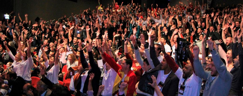 A large audience on their feet with arms raised during a Fabio Marques keynote