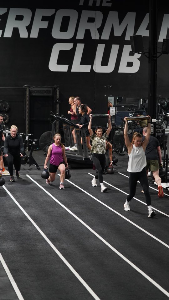 Members lunging up the track with kettlebells and plates overhead under the club sign