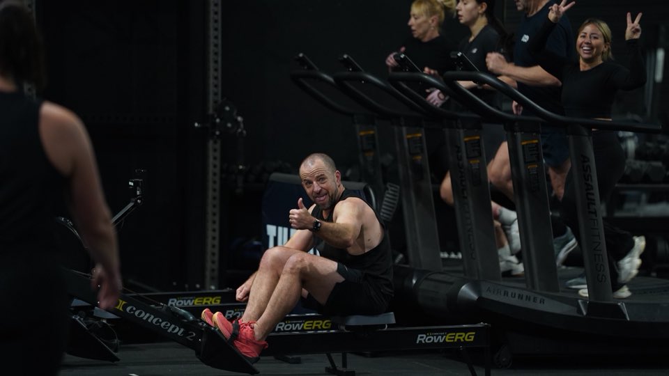 A member on the rower grinning with a thumbs-up while a classmate waves from the treadmill
