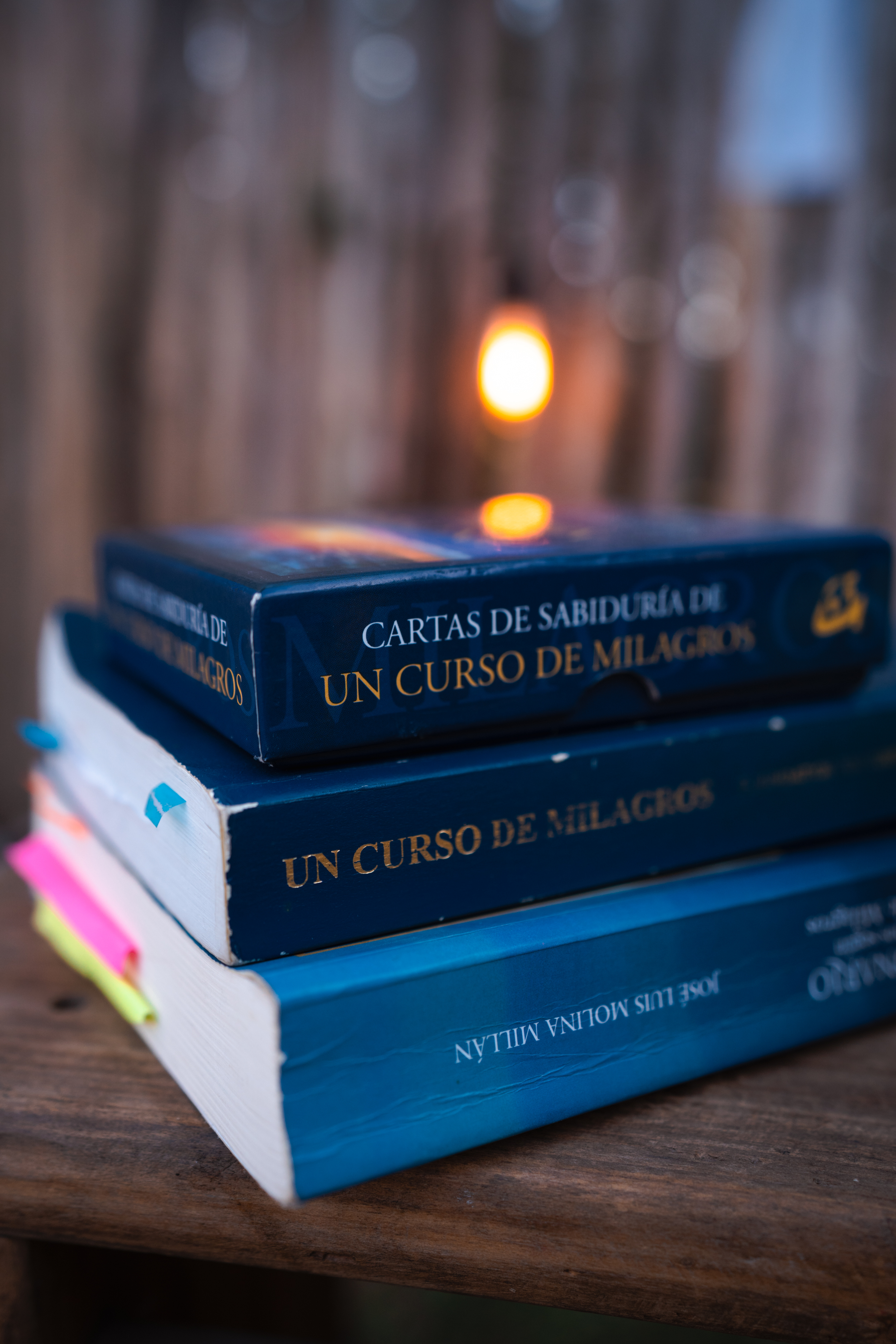 Un Curso de Milagros, sus cartas de sabiduría y otro libro apilados sobre una mesa de madera, con una luz cálida detrás