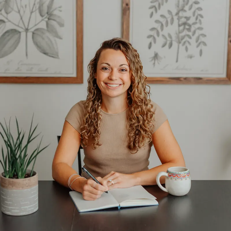Jen Navon smiling at her desk with a pen and open notebook, botanical prints on the wall behind her