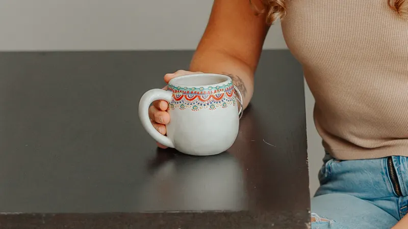 Jen's hand wrapped around a painted coffee mug on a dark wooden table