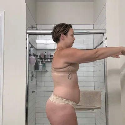 Jen in 2025, a side-view postpartum progress photo in her bathroom with her arms held out
