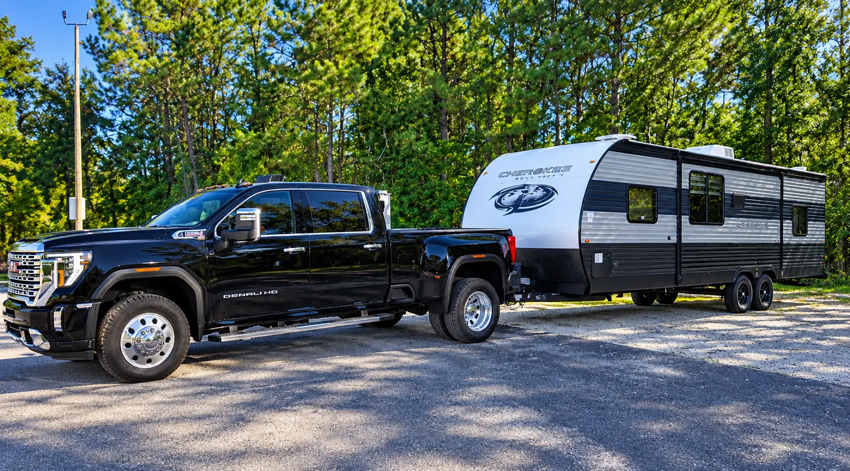 Black GMC Denali HD truck towing a Cherokee travel trailer beside pine trees.