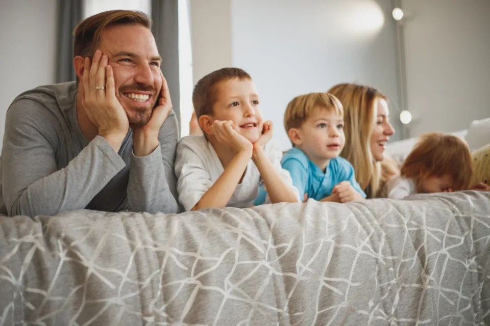 Dad and kids grinning together on a bed in a calm, freshly painted bedroom