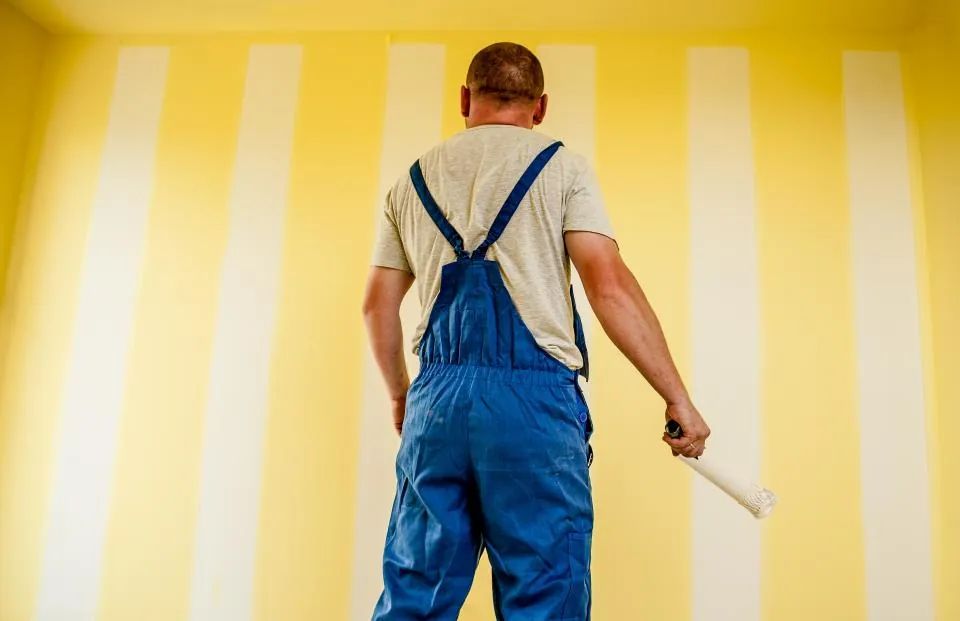 Painter in overalls with a roller, standing before a freshly painted yellow and white striped wall