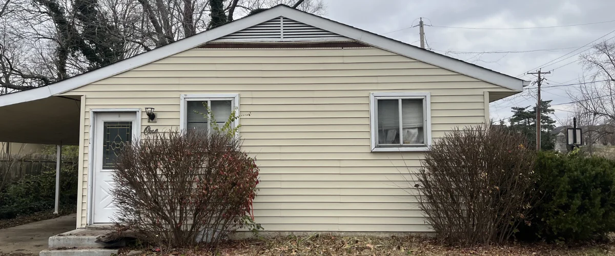 A section of a house with pale yellow siding, a white-framed window with blinds, and green bushes below.