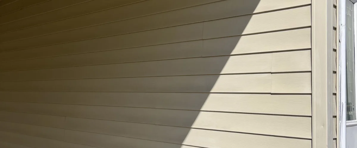A tan house siding with horizontal panels, partially lit by sunlight with a diagonal shadow across it.