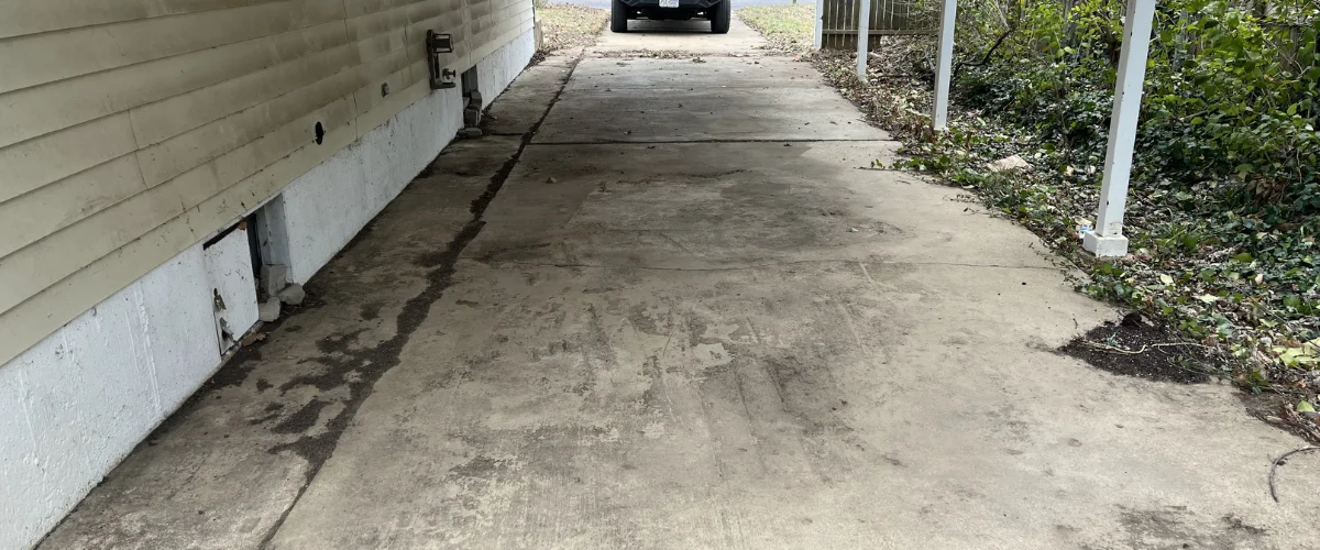 A long, narrow concrete driveway next to the side of a house with light yellow siding and a car parked in the distance. A long, narrow concrete driveway next to the side of a house with light yellow siding and a car parked in the distance.