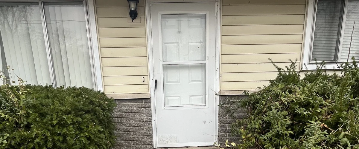 A weather-beaten white storm door on a house with horizontal yellow siding, surrounded by green shrubs. A weather-beaten white storm door on a house with horizontal yellow siding, surrounded by green shrubs.