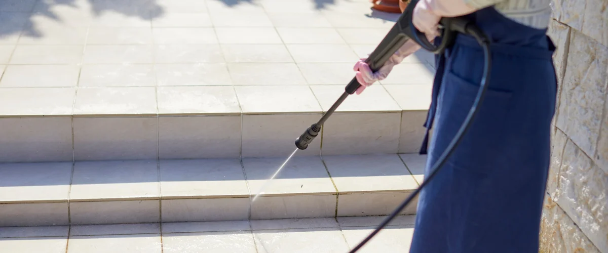 A person in a blue apron and pink gloves uses a pressure washer to clean light-colored patio steps.