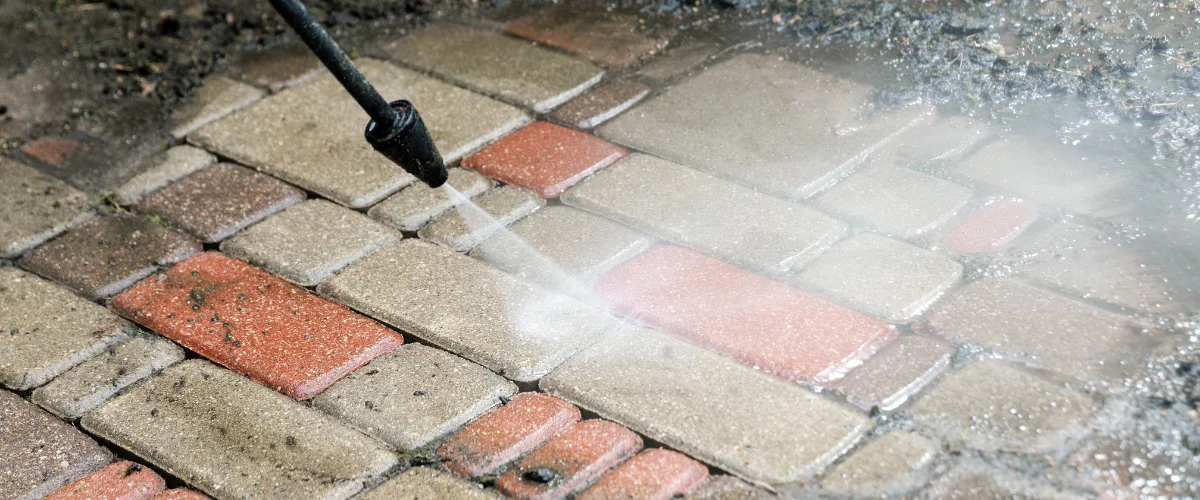 Close-up of a pressure washer cleaning dirty red and gray patio pavers, showing water spray and clean sections.