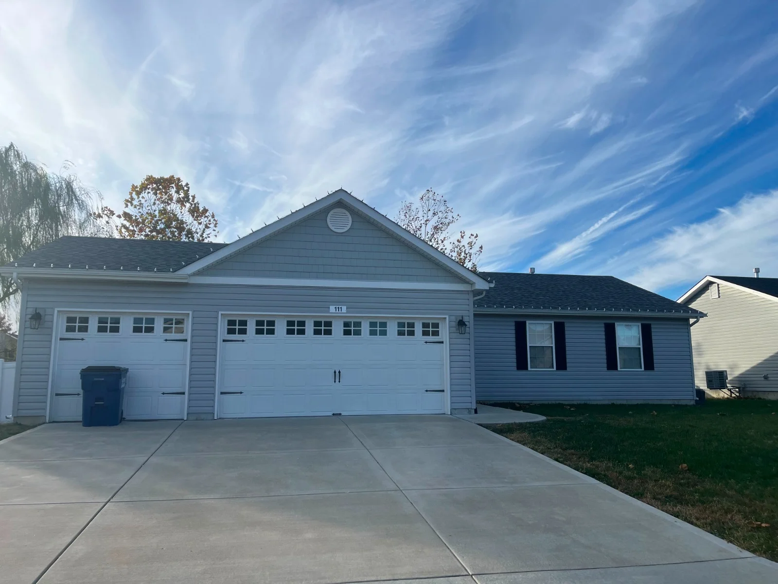 A light gray house with two white garage doors and black shutters, a concrete driveway, and wispy clouds in the blue sky.
