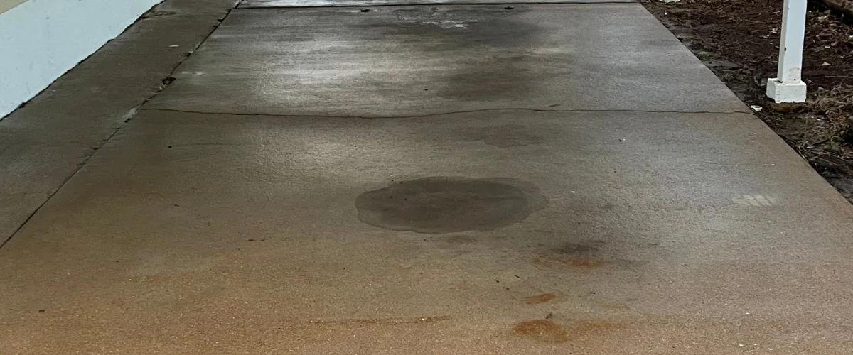 A concrete sidewalk with wet spots and small puddles, bordered by a white wall on the left and dirt on the right.