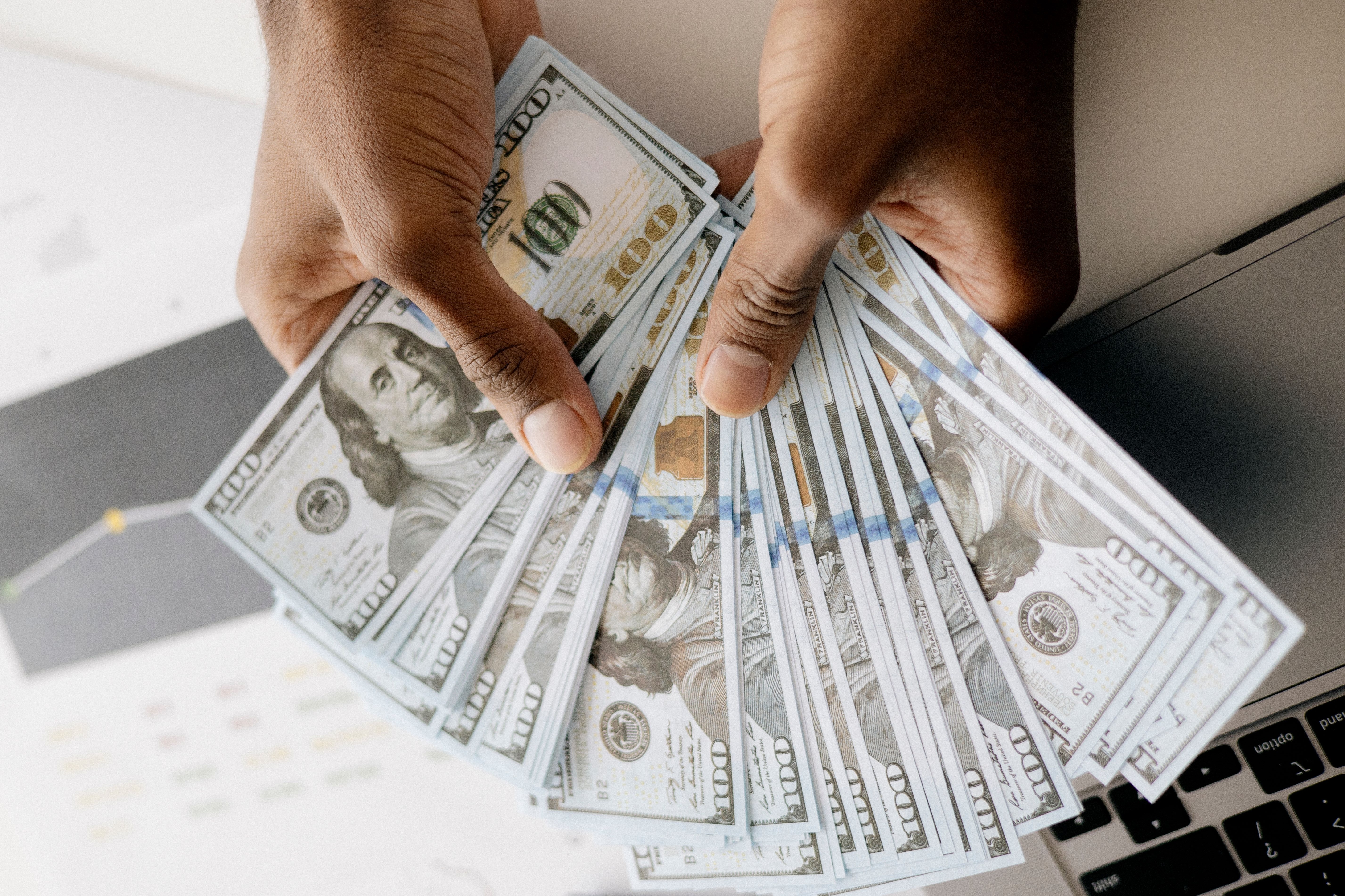 Hands fanning out a stack of hundred-dollar bills above a laptop