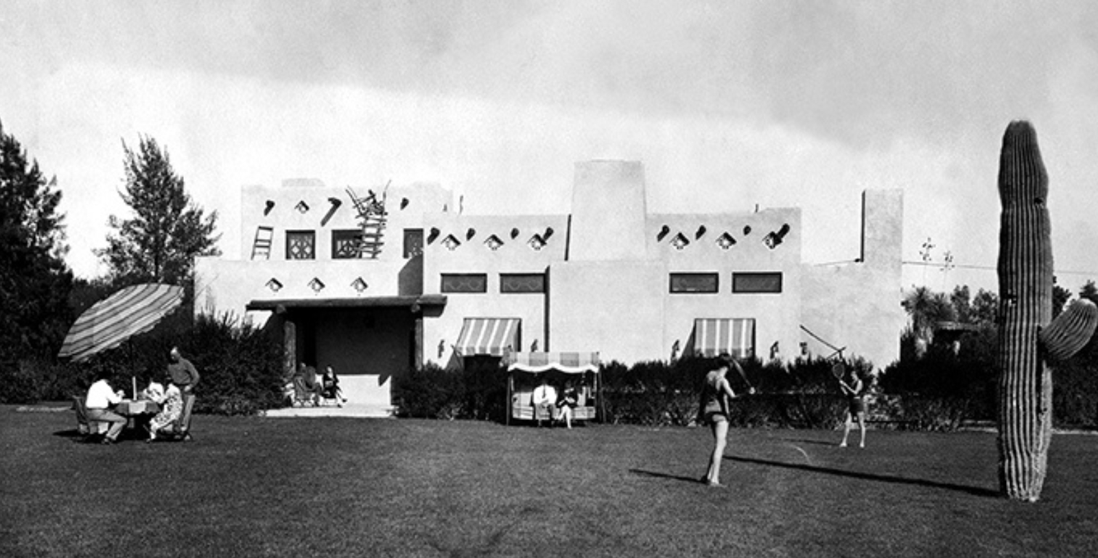 Historic black-and-white photograph of The Wigwam Resort in Litchfield Park, Arizona — showing Pueblo Revival adobe architecture, striped awnings, desert landscaping, saguaro cactus, and guests relaxing on the lawn