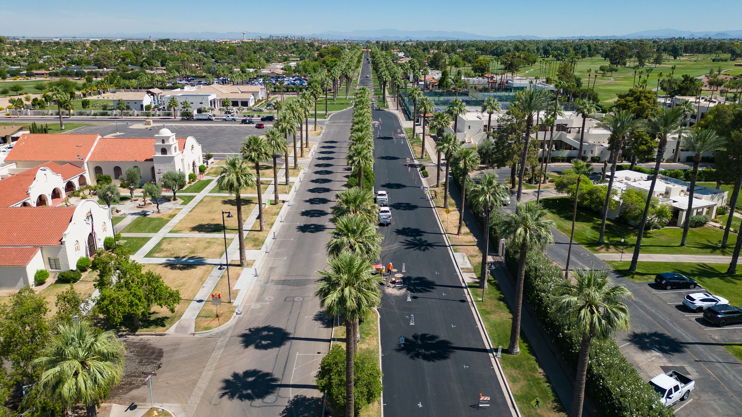 Contemporary aerial photograph of Old Litchfield Park, Arizona — showing the long palm-lined boulevard, Spanish Colonial Revival buildings with red-tile roofs, tennis courts, lush green lawns, and the resort-town layout unchanged from its Goodyear-era origins