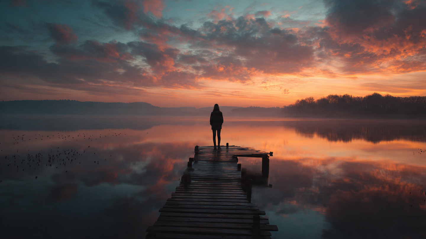 A person standing outdoors at sunrise on, The end of a pier overlooking a calm lake, posture upright and calm, hands at sides, confident but peaceful expression. Birds trees in the distance blurred beautiful multi colored clouds in the sky.