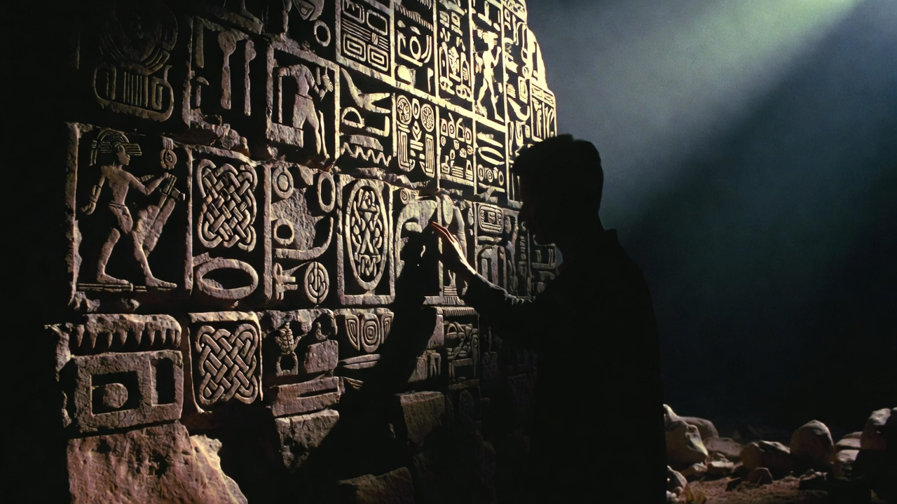 A person standing before a massive ancient stone wall covered in carved symbols and hieroglyphics from multiple cultures (Egyptian, Mayan, Celtic) blending together. The person is touching the wall gently, fingers tracing the carvings, shaft of light illuminating their hand. A person standing before a massive ancient stone wall covered in carved symbols and hieroglyphics from multiple cultures (Egyptian, Mayan, Celtic) blending together. The person is touching the wall gently, fingers tracing the carvings, shaft of light illuminating their hand.
