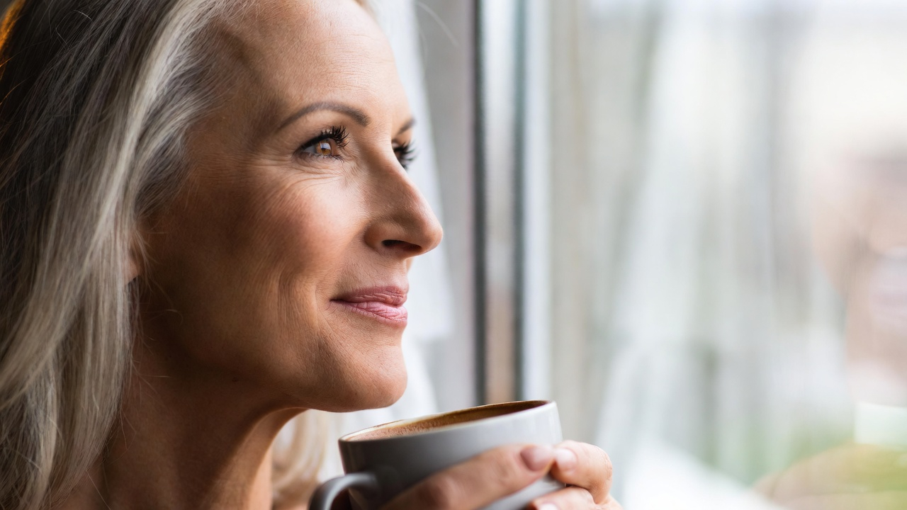 An image of a woman in her 50s smiling gently while looking out a window. She is holding a warm mug. The focus is on her eyes, which look clear, bright, and peaceful. The image should convey a sense of regained mental clarity and emotional resilience. Soft, natural lighting. An image of a woman in her 50s smiling gently while looking out a window. She is holding a warm mug. The focus is on her eyes, which look clear, bright, and peaceful. The image should convey a sense of regained mental clarity and emotional resilience. Soft, natural lighting.