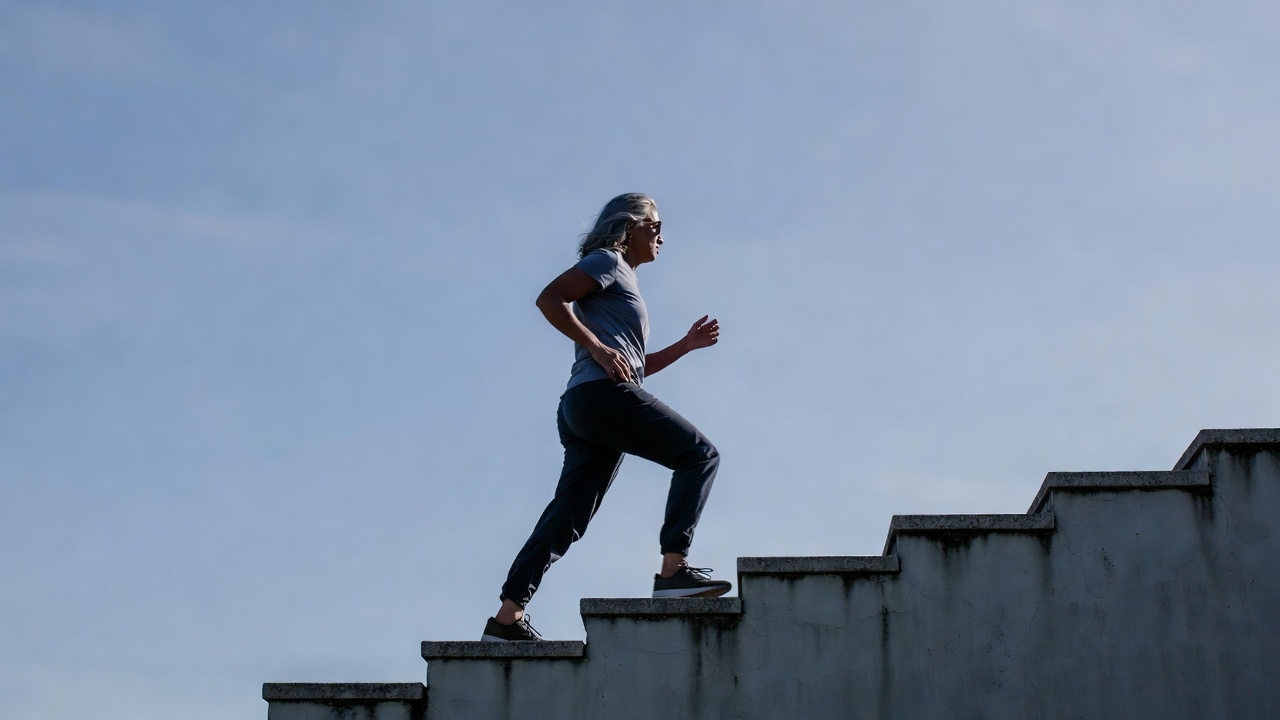 A dynamic, slightly motivational photo of a person (diverse, age 40-60) walking confidently up a set of stairs or a hill, looking forward with purpose. The path is ahead of them, the sky is clear. They are not struggling, but moving with steady, renewed strength. It symbolizes moving forward into the future with power. A dynamic, slightly motivational photo of a person (diverse, age 40-60) walking confidently up a set of stairs or a hill, looking forward with purpose. The path is ahead of them, the sky is clear. They are not struggling, but moving with steady, renewed strength. It symbolizes moving forward into the future with power.