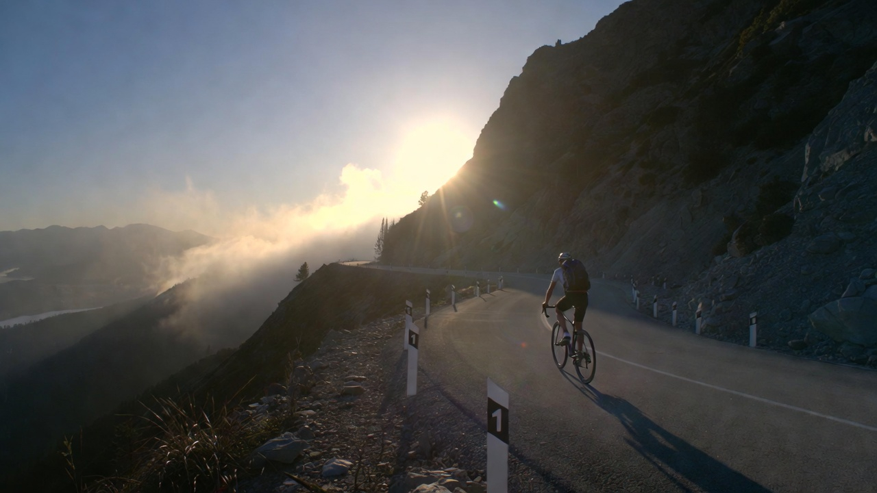 A cyclist climbing a steep mountain road at dawn. The road winds upward with tiny, almost invisible incremental markers along the edge representing 1% gains. The cyclist is small in the frame, emphasizing the magnitude of the journey. Soft morning light, mist in the valleys.