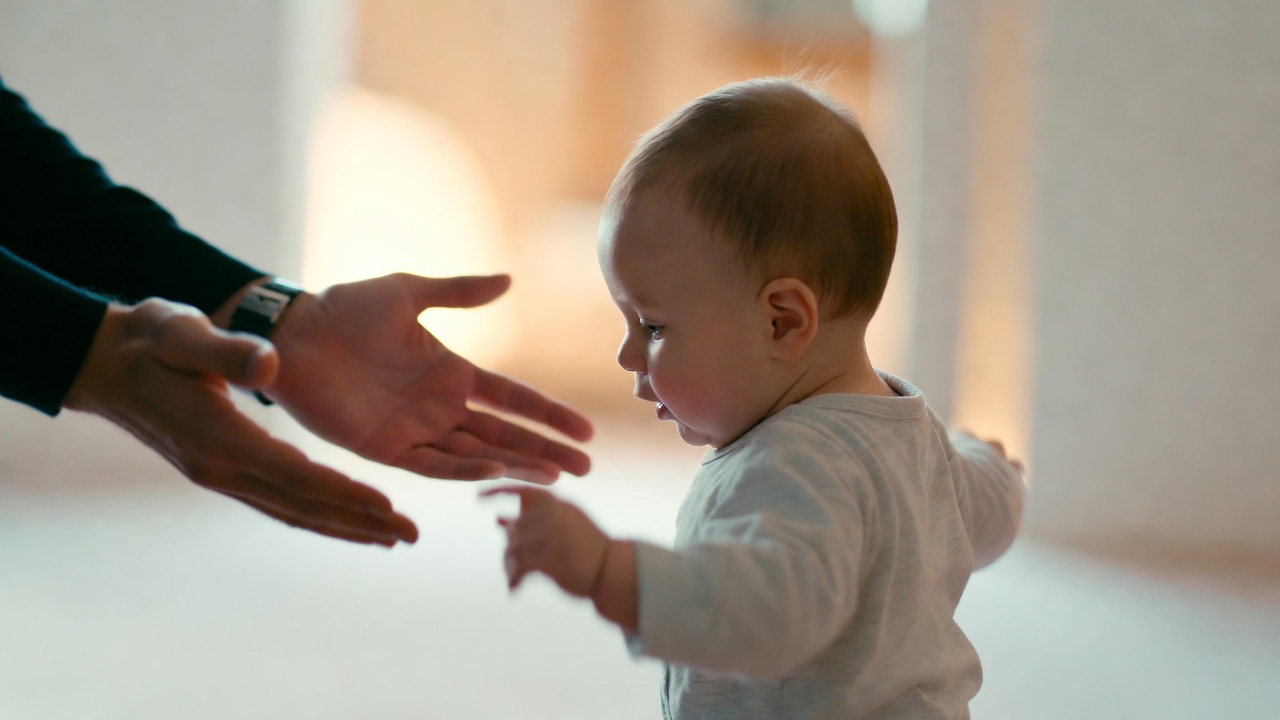 A baby taking their first steps toward a parent's outstretched hands. Soft, warm lighting. The background is slightly blurred, emphasizing the moment of determination and the support waiting ahead