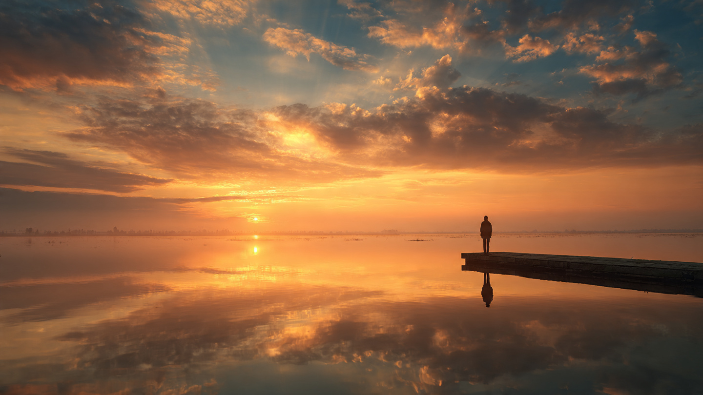 Sunrise over calm horizon Silhouette of lone dark figure at the end of a pier enjoying the beautiful sunrise over the lake