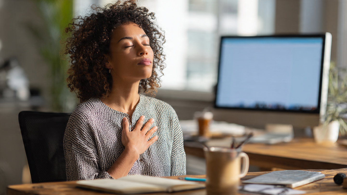 A person sitting at their desk in the middle of a busy day. Eyes closed. One hand on their chest, one on their lap. Taking three deep breaths. Soft natural light from a window. In front of them on the desk: a notebook with a pen resting on it, a coffee mug, and a single open book. Behind them, slightly blurred, a computer screen shows work—emails, calendar, tasks. The contrast is clear: the world is waiting, but they chose this moment. The mood is calm, centered, defiantly peaceful A person sitting at their desk in the middle of a busy day. Eyes closed. One hand on their chest, one on their lap. Taking three deep breaths. Soft natural light from a window. In front of them on the desk: a notebook with a pen resting on it, a coffee mug, and a single open book. Behind them, slightly blurred, a computer screen shows work—emails, calendar, tasks. The contrast is clear: the world is waiting, but they chose this moment. The mood is calm, centered, defiantly peaceful