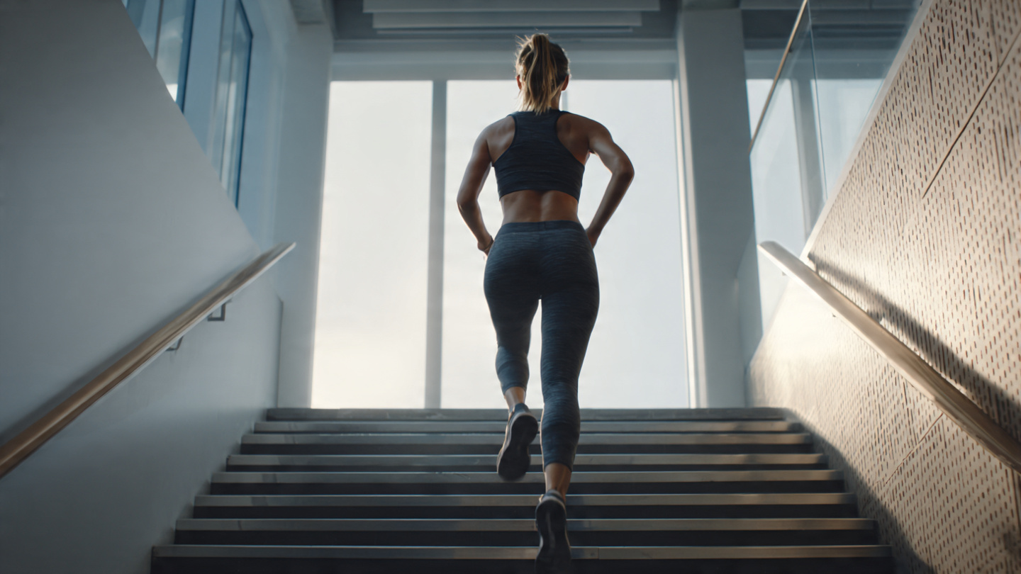 A single woman, age 30s, standing at the bottom of a staircase in a modern gym or office building, mid-motion about to sprint up the stairs. She's wearing athletic wear (leggings and tank top), her face shows determination, one foot on the first step
