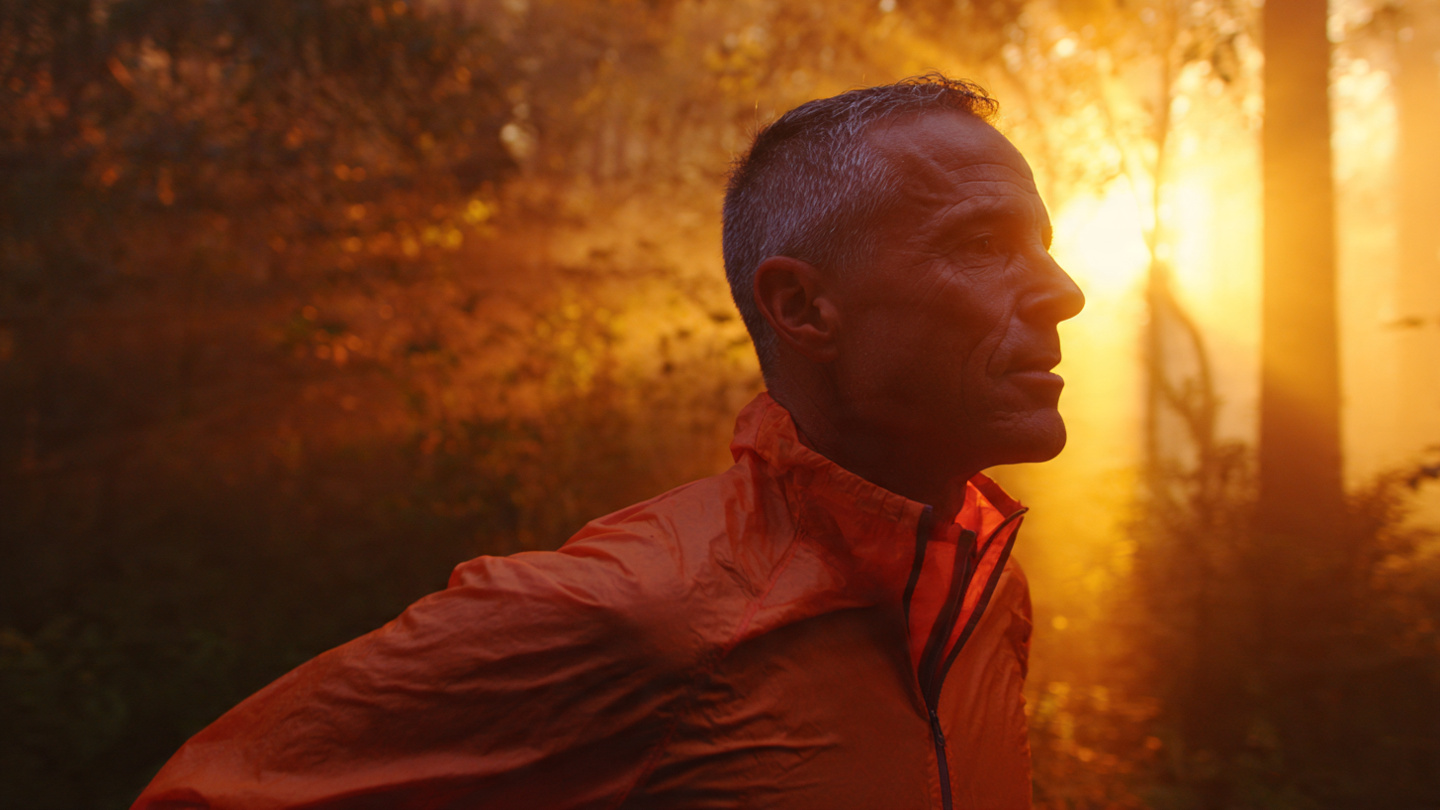 A strong, vibrant man in their late 40s or 50s jogging on a scenic trail at sunrise — golden light cutting through trees, breath visible in the cool morning air. Radiating energy, vitality, and purpose. The image should feel like the result of doing everything right. Inspirational, cinematic, aspirational health and longevity aesthetic.