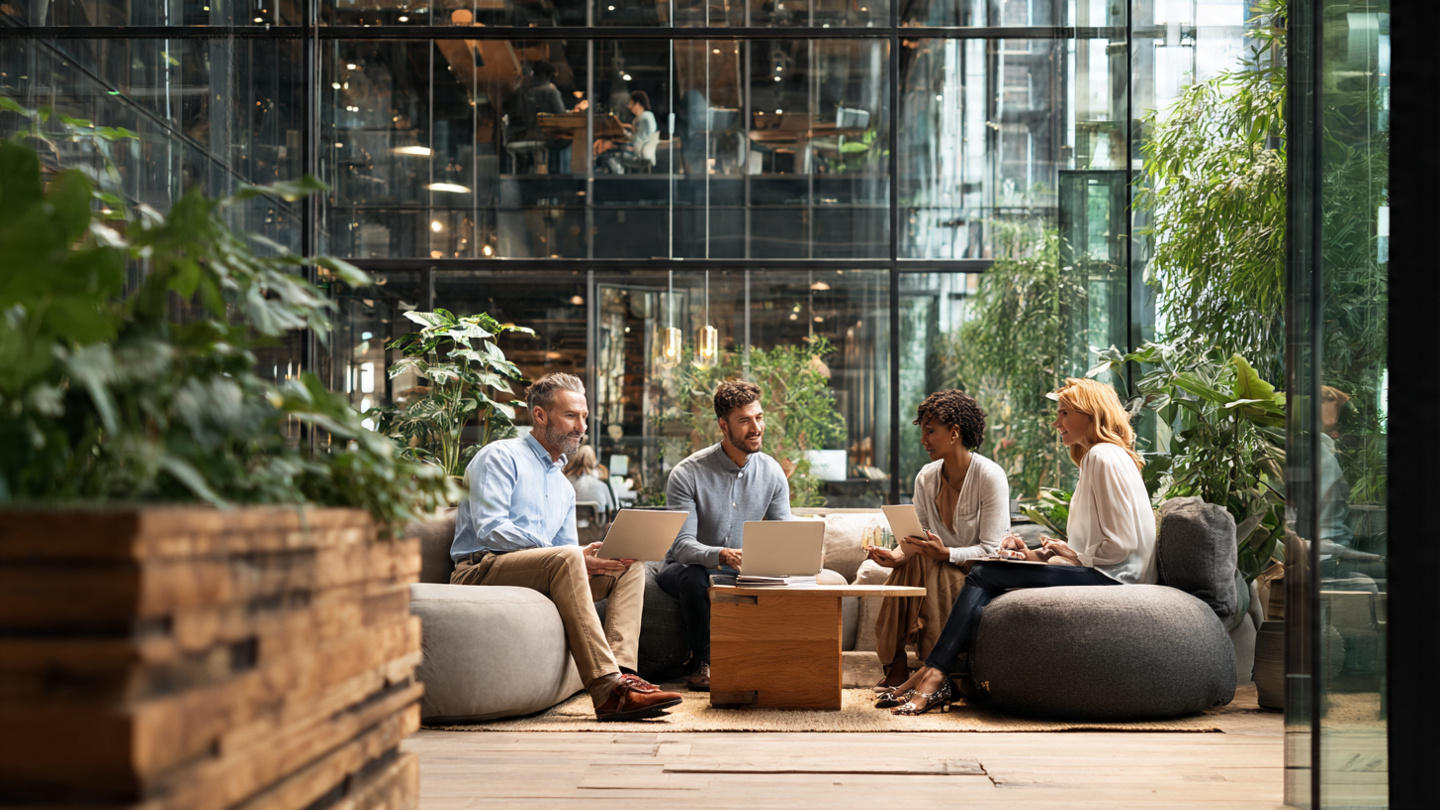 A diverse group of 4 professionals in a modern collaborative workspace, looking at tablets and laptops together, engaged in discussion, natural lighting, contemporary office design with glass walls and plants